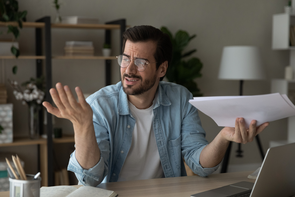 frustrated young man in eyewear holding paper correspondence in hands
