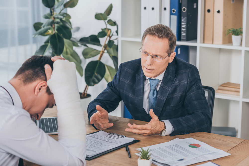 worker with broken arm sitting at table and reading documents opposite to businessman in blue jacket in office