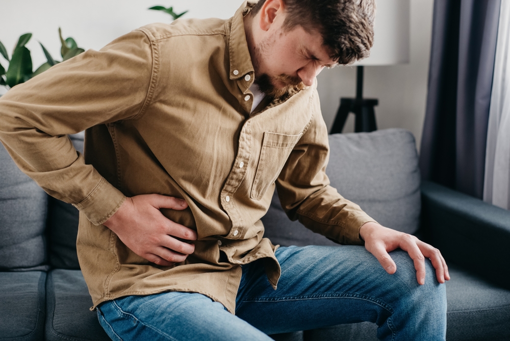 Unhealthy young caucasian man 30s holding belly sit on couch at home feel bad digestive problems abdominal pain illness suffers from liver damage bending body in ache feeling unwell poisoning